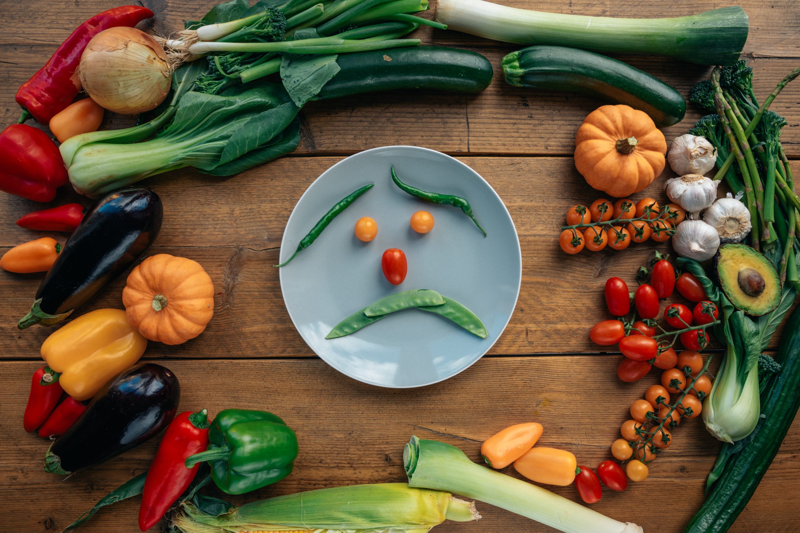 Une assiette avant des légumes positionnés en forme de tete triste, entourée de fruits et légumes, posée sur une table en bois